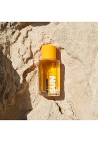 Gjennomsiktig rektangulær glassflaske fylt med gul væske, toppet med en lys gul kork. Hvit etikett med blå tekst: "JIL SANDER SUN."