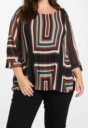 Vrouw die een meerkleurige gestreepte blouse met 3/4 mouwen en zwarte broek draagt, staand tegen een effen witte achtergrond.