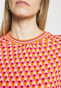 Nainen, jolla on suorat vaaleat hiukset, käyttää kultaisia rengassolia ja värikästä kuvioitua paitaa, jossa on oransseja, vaaleanpunaisia ja punaisia soikioita.