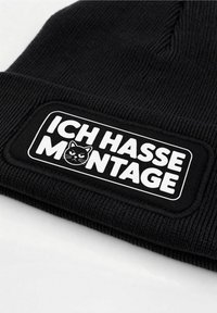 Black knit beanie featuring a rectangular patch with the text "ICH HASSE MONTAGE" and a cat graphic. Text is white on a black background.
