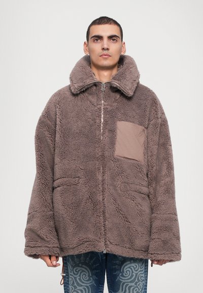 Chaqueta de forro polar marrón con un cuello alto, cierre de cremallera y un bolsillo de parche en la parte frontal. Cuenta con una superficie suave y texturada y un corte oversize.