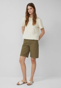 Korte mouwen crème t-shirt, olijfgroene opgerolde shorts en witte platte sandalen. Eenvoudig ontwerp, zacht materiaal, neutrale kleuren.