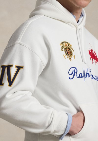 Osoba ubrana w białą bluzę z kapturem z niebieskim napisem "Ralph Lauren", czerwonym logo polo gracza, złotym herbem oraz dużą niebieską rzymską cyfrą IV na rękawie.