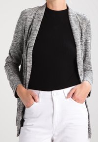 Lehký šedý kardigan s dlouhým rukávem a otevřeným předním dílem s jemnou strukturou, kombinovaný s černým žebrovaným topem a bílými kalhotami.