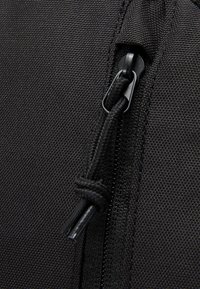 Detail van een zwarte stoffen tas met een ritsnaad, textuur nylon en een trekkoord met een plastic punt. De stiksels zijn zichtbaar en strak.