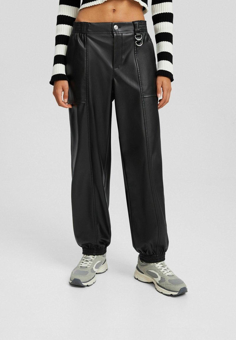 Personne portant un pantalon jogger en simili cuir noir avec des poignets élastiques, associé à des baskets grises et blanches et un pull court rayé noir et blanc.