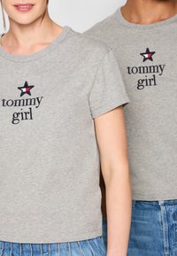 Pilkos medvilninės marškinėliai su trumpomis rankovėmis, puošti žvaigžde ir juodu siuvinėjimu „tommy girl“ su raudonais ir baltais akcentais.