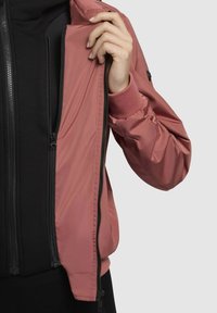 Veste bomber rose pâle avec fermeture éclair sur le devant, texture douce, poignets côtelés et une couche intérieure noire contrastante visible à la fermeture éclair ouverte.