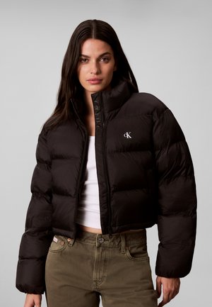 Vrouw draagt een zwart cropped pufferjack met een witte top en olijfgroene broek, staand tegen een effen grijze achtergrond.