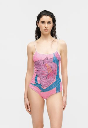 Femeie purtând un costum de baie întreg roz, cu un model mare de flori mov și albastre, stând în picioare pe un fundal simplu, deschis la culoare.