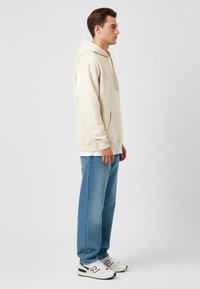 Hoodie beige con tasca frontale, vestibilità rilassata, abbinato a jeans baggy azzurri e sneakers bianche con accenti neri.