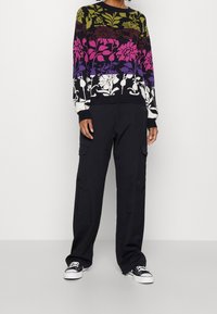Personne portant un pantalon cargo noir, des baskets noires avec des semelles blanches, et un pull floral multicolore avec des motifs verts, violets, roses et blancs.