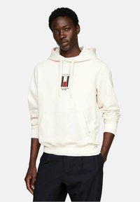 Sudadera con capucha color crema, con parche de logo en el frente, cuello con cordones y puños acanalados. Combinada con pantalones oscuros. Textura de tela suave.