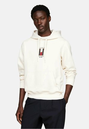 Sudadera con capucha color crema, con parche de logo en el frente, cuello con cordones y puños acanalados. Combinada con pantalones oscuros. Textura de tela suave.