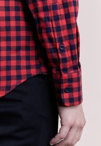 Brazo que lleva una camisa de manga larga a cuadros rojos y negros con botones marinos, descansando al lado de pantalones negros sobre un fondo claro y liso.