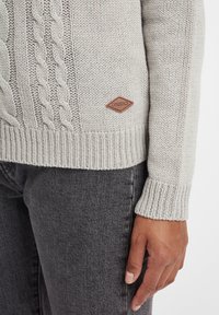 Maglione lavorato a maglia grigio chiaro con motivi a treccia, polsini a costine e un'etichetta in pelle sul lato. Indossato con jeans grigio scuro.