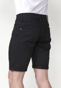 Muž v čiernych džínsových šortkách s ohrnutými okrajmi a bielou košeľou, zobrazený zo zadného pohľadu od pásu po stred stehien.