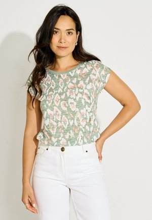 Mujer con cabello largo y oscuro, vistiendo una blusa verde de mangas cortas con estampado y pantalones blancos, de pie con una mano en la cadera sobre un fondo blanco.
