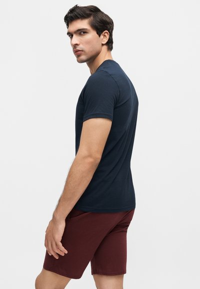 Άνδρας που στέκεται πλευρικά κοιτώντας πάνω από τον ώμο, φοράει μπλε σκούρο κοντομάνικο μπλουζάκι και μπορντό σορτς σε απλό ανοιχτό φόντο.