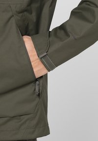 Olivengrøn vandtæt jakke med lynlåslomme til hænder, tekstureret stof og justerbar manchet med velcrolukning.