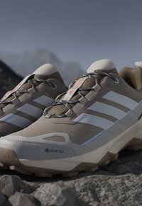 Chaussures de randonnée beiges et blanches avec semelles Gore-Tex et Lightmotion posées sur un terrain rocheux avec des montagnes en arrière-plan.