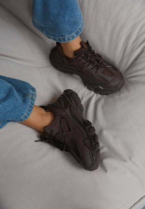 Füße in dunkelbraunen, klobigen Sneakers und blauen Jeans ruhen auf einer hellgrauen Stoffoberfläche.