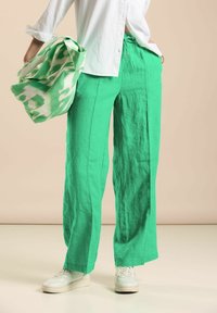Green linen wide-leg pants with a drawstring waist, paired with a white button-up shirt and a patterned green and white bag.