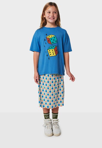 Blaues Grafik-T-Shirt mit Würfel-Motiv, kombiniert mit einem plissierten Rock, der mehrfarbige Punkte auf weißem Hintergrund zeigt. Weiße Sneakers und grüne Socken.