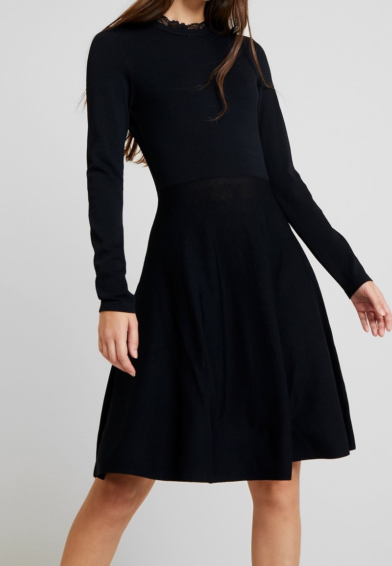 Femme portant une robe noire mi-longue à manches longues avec une jupe légèrement évasée et un détail en dentelle sur le décolleté, sur un fond uni.