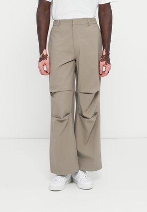 Pantaloni a gamba larga di un marrone chiaro, realizzati in un tessuto liscio con pieghe frontali; indossati con una camicia bianca e sneakers bianche.