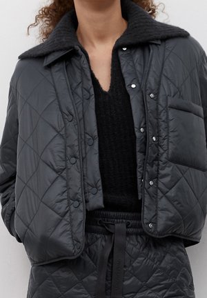 Kvinde iført sort quiltet jakke med trykknapper, stor krave og matchende quiltede bukser med snøre i taljen.