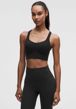 Vrouw met donker gevlochten haar in een zwarte sport-bh en hoge taille legging, staand tegen een effen lichte achtergrond.