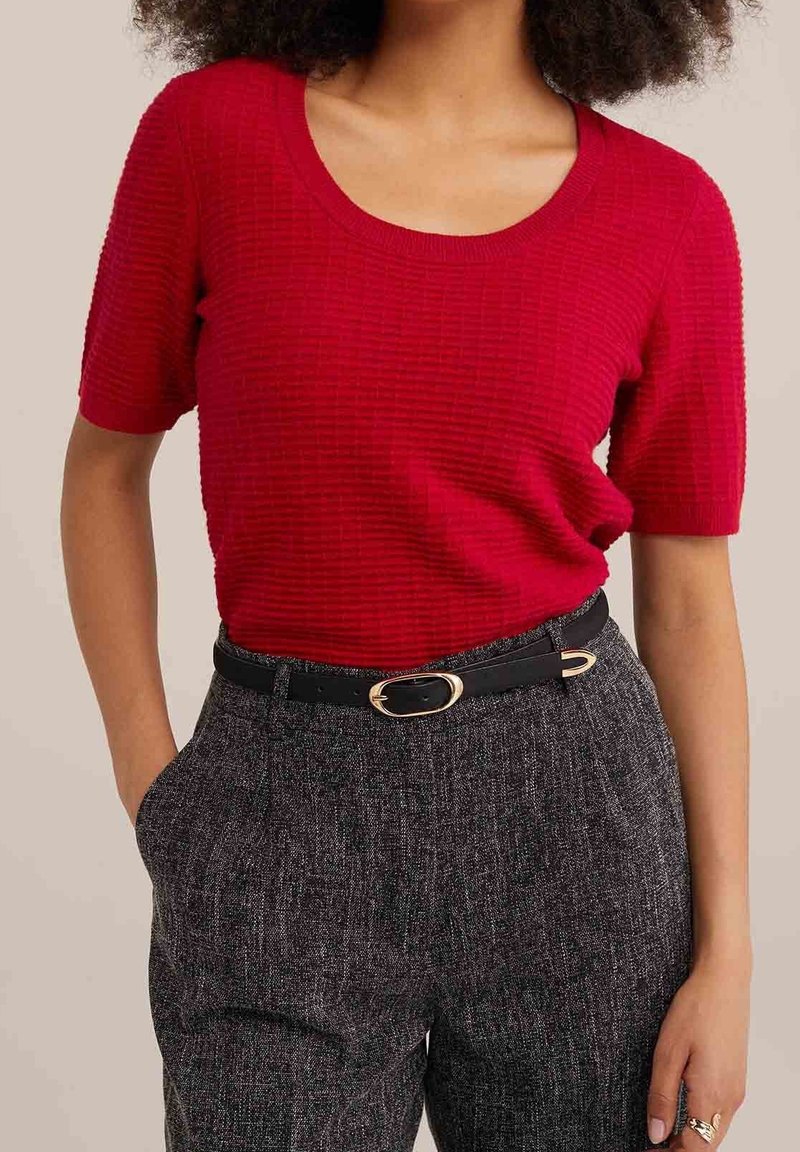 Haut en tricot rouge côtelé avec un col rond et des manches courtes, associé à un pantalon gris texturé et une ceinture noire ornée d'une boucle ovale dorée.