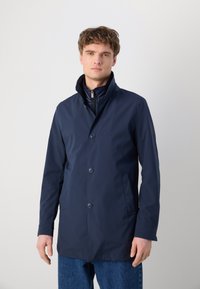 Giovane uomo che indossa un cappotto blu navy con bottoni e colletto alto sopra uno strato interno con zip, in piedi su uno sfondo chiaro e uniforme.