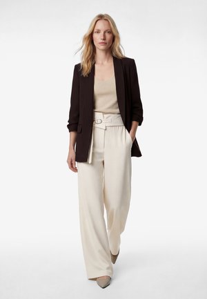 Blonde Frau geht nach vorne und trägt einen schwarzen Blazer, ein beiges Oberteil, weiße hoch taillierte weite Hosen mit Gürtel und beigen spitzen Schuhen.