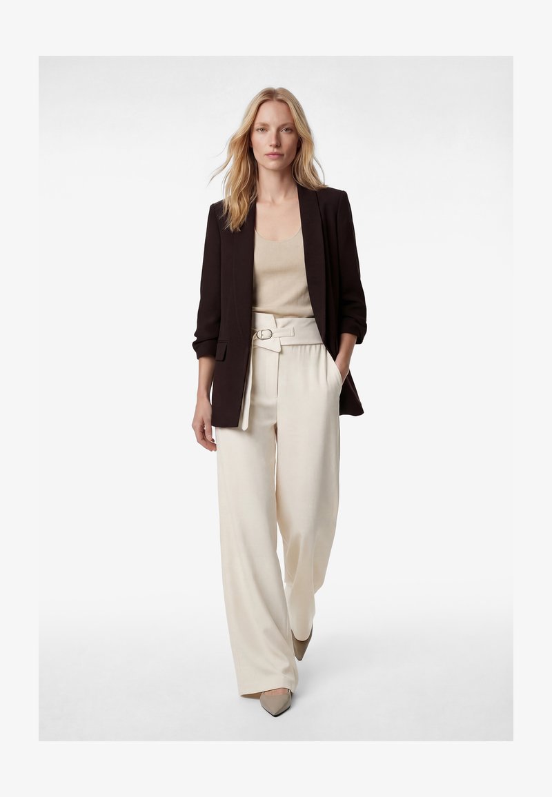 Mujer rubia caminando hacia adelante, vestida con blazer negro, top beige, pantalones blancos de pierna ancha y cintura alta con cinturón, y zapatos beige puntiagudos.