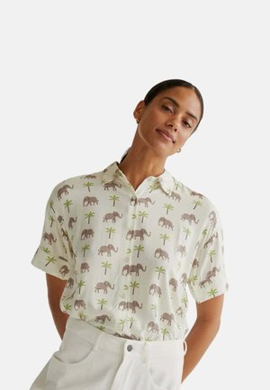 Mujer que lleva una camisa blanca de manga corta con botones y un patrón de pequeños elefantes y palmeras verdes, combinada con pantalones blancos.