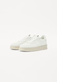 Baskets en cuir blanc avec une surface texturée, bout rond, lacets et épaisse semelle en caoutchouc crème. Marquage subtil sur le côté.
