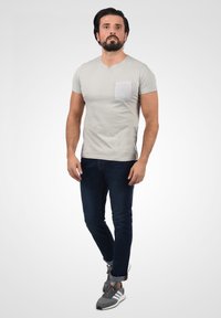 Svijetlo siva majica kratkih rukava s v-izrezom i punom džepom, u kombinaciji s tamnoplavim slim-fit trapericama i sivim sportskim cipelama.