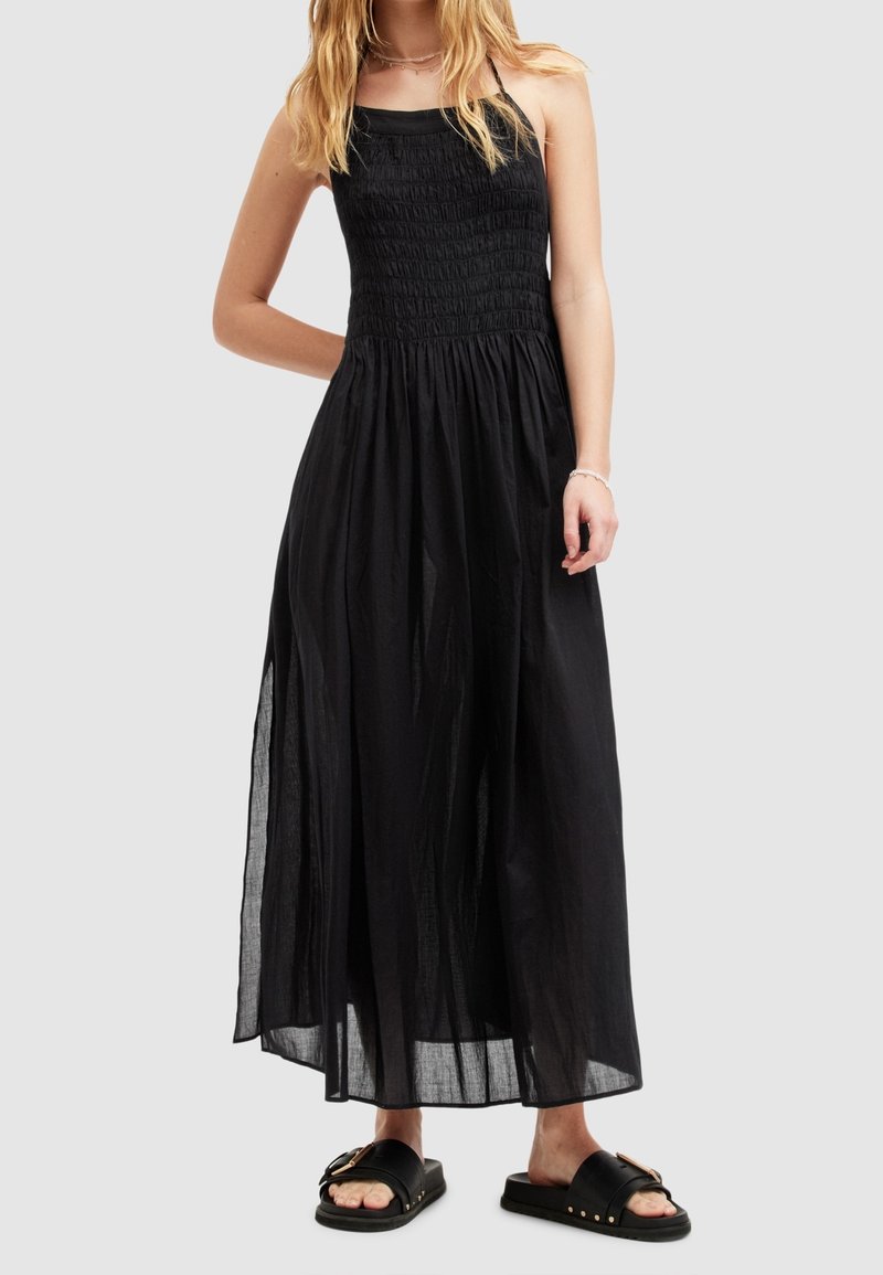 Femme portant une robe maxi noire sans manches avec un corsage smocké et des sandales slides noires avec des accents de boucle dorée.