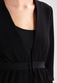 Zwarte cardigan met driekwart mouwen, plooien aan de voorkant en een gladde riem in de taille. Zachte stof met een subtiele val.