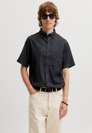 Joven con camisa negra de manga corta abotonada, pantalones beige, cinturón negro, gafas de sol y una pulsera negra de cuentas, de pie con una mano en el bolsillo.