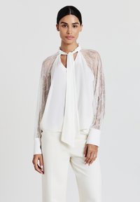 Donna che indossa una blouse bianca con maniche in pizzo e colletto a cravatta, abbinata a pantaloni bianchi su uno sfondo chiaro e uniforme.
