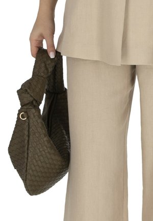 Dłoń trzyma ciemnobrązową, plecioną skórzaną torbę z węzłowym uchwytem, zestawioną z beżowymi lnianymi spodniami i górą.