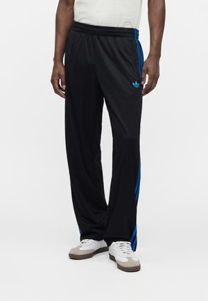 Άντρας που φοράει μαύρο παντελόνι φόρμας Adidas με μπλε ρίγες και λογότυπο, λευκά αθλητικά παπούτσια και λευκό πουκάμισο σε απλό φόντο.
