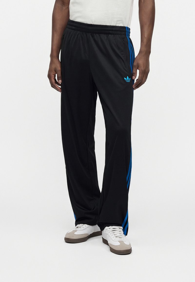 Άντρας που φοράει μαύρο παντελόνι φόρμας Adidas με μπλε ρίγες και λογότυπο, λευκά αθλητικά παπούτσια και λευκό πουκάμισο σε απλό φόντο.
