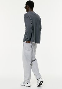 Camicia a maniche lunghe grigia con vestibilità comoda, abbinata a jeans larghi grigi chiari. Sneakers bianche con dettagli neri. Design minimalista.