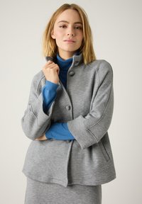 Grijze, zachte stoffen jas met een hoge kraag en drie knopen, gecombineerd met een blauwe coltrui. Heeft een ontspannen pasvorm en driekwart mouwen.