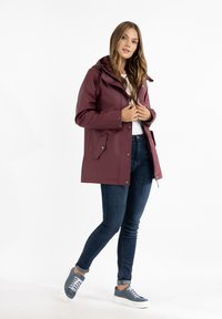 Burgundy regnfrakke med hætte, trykknap- og lynlåslukning, to sidelommer, båret over en hvid skjorte og mørke denimjeans med grå sneakers.