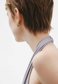 Aro de oro en una persona con el cabello corto, con una tela gris claro que cae en el área del cuello y los hombros. Textura de piel suave visible.